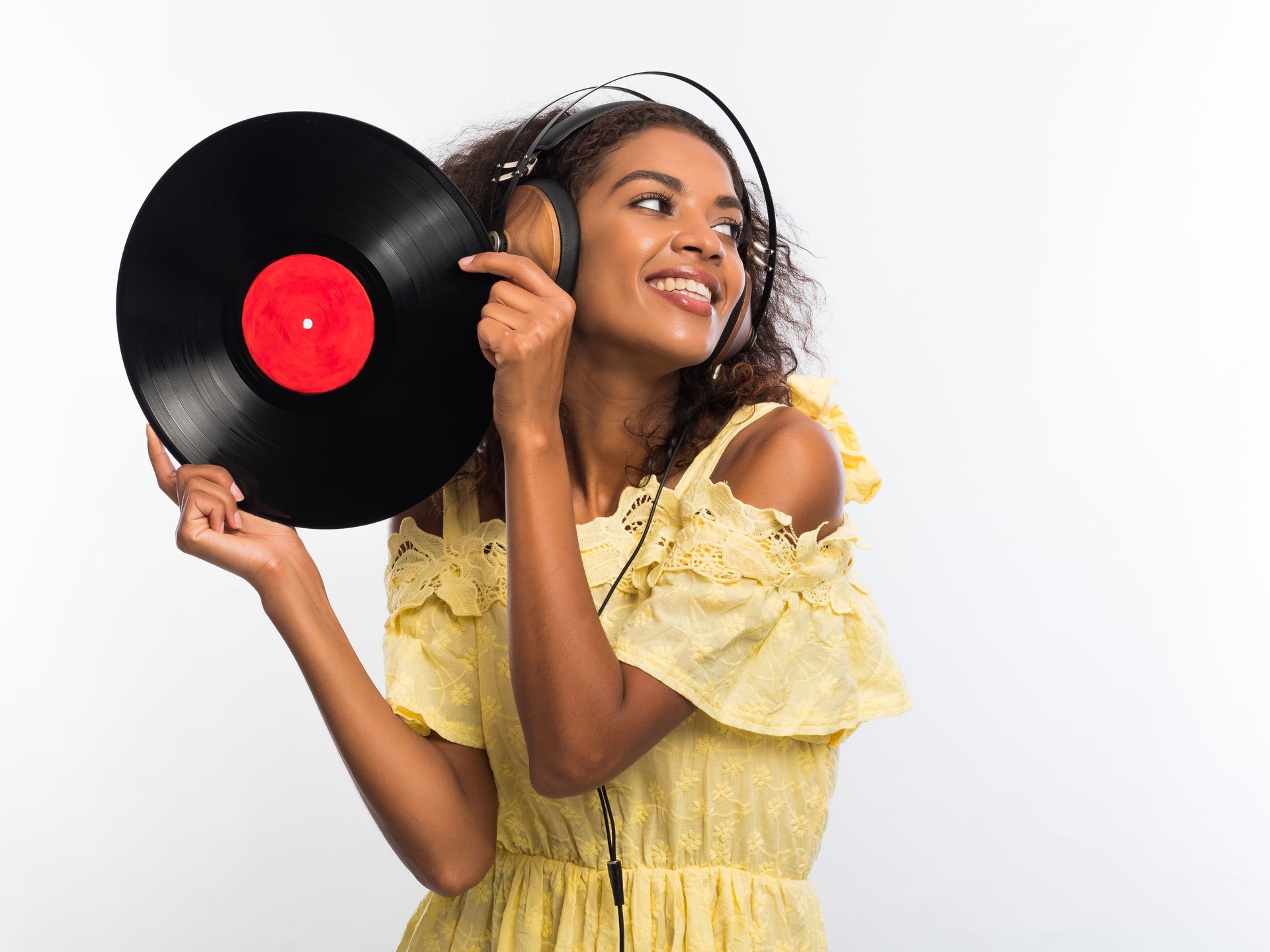 Young beautiful african american woman with headphones in yellow dress enjoying and dancing with vinyl records at white background.